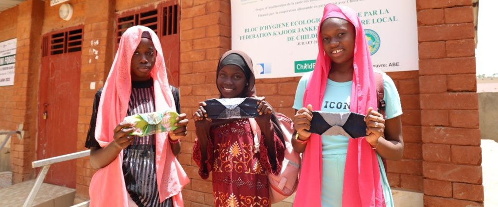 Fatou, 13 ans, Zainab, 14 ans, et Mame, 14 ans, tenant une serviette hygiénique réutilisable à l'extérieur du bloc sanitaire de leur collège